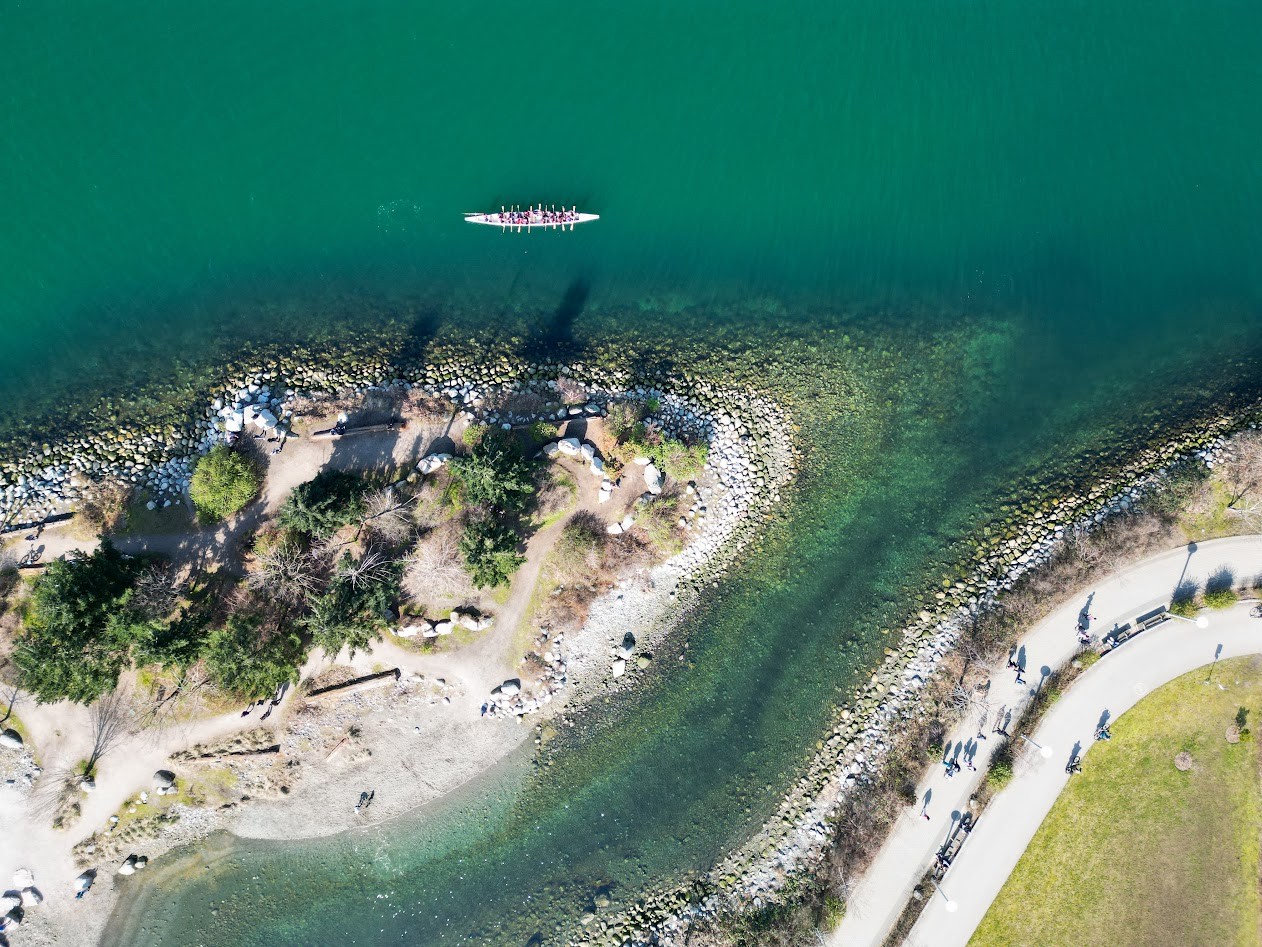 An aerial view of a rowing crew on green water beside a waterfront path